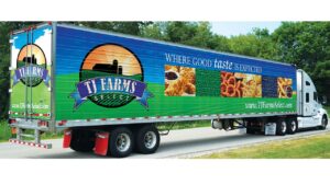 A TJ Farms Select truck with food images on the side and the slogan "Where good taste is expected" on a road with trees in the background.