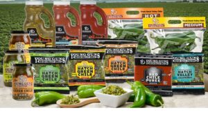 Various Young Guns brand chile products are displayed, including jars, sauces, and packaged peppers, surrounded by fresh green chiles. A field is visible in the background.